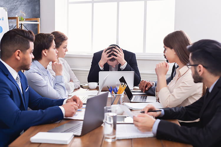 Photo of 7 people in a meeting, the meeting leader shows frustration with hands on head